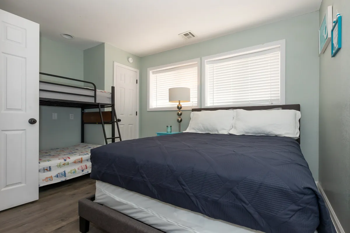 A family-friendly bedroom scene with a bunk bed and a main bed, softly illuminated by a lamp and natural window light. The space is designed for relaxation, reading, and family bonding, ideal for a variety of group arrangements.