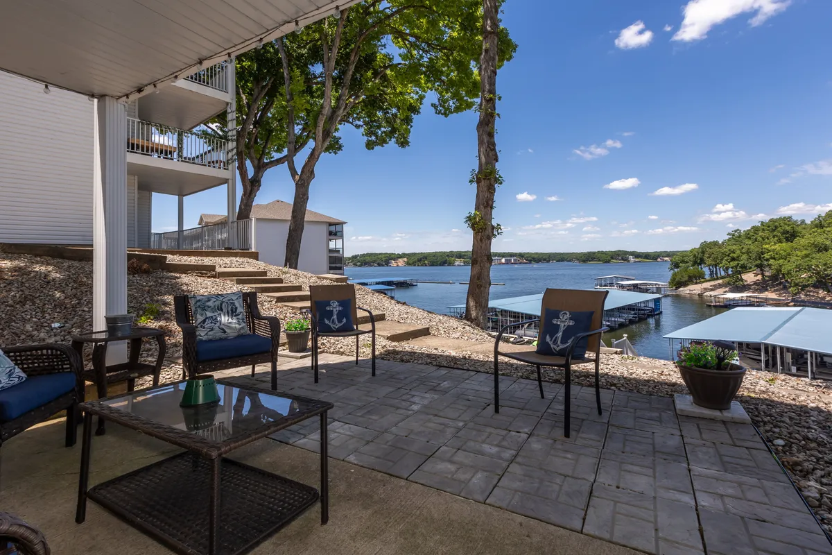 The image showcases a tranquil lakefront outdoor seating area at a pet-friendly condo in Osage Beach, Missouri. The view includes sparkling water, lush decorative plants, and a cozy spot to relax and enjoy the passing boats, all just steps from the water's edge.