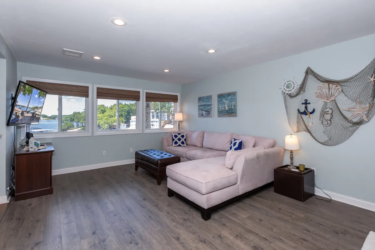 A comfortable living room with a plush sofa, television, windows letting in natural light, and a decorative wall hanging. The arrangement suggests relaxing evenings, movie nights, or simply enjoying the view outside.