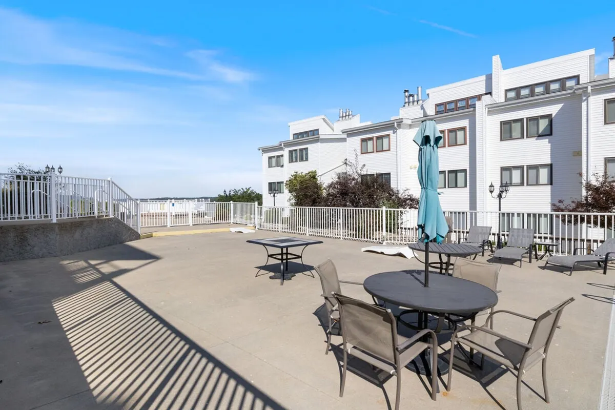 An exterior shot of a fenced patio area, furnished with tables, chairs, and an umbrella, designed for comfort and socializing. The fenced space hints at safety for pets, while the relaxed atmosphere is ideal for morning coffees, sunset views, or spending time outdoors with animals.