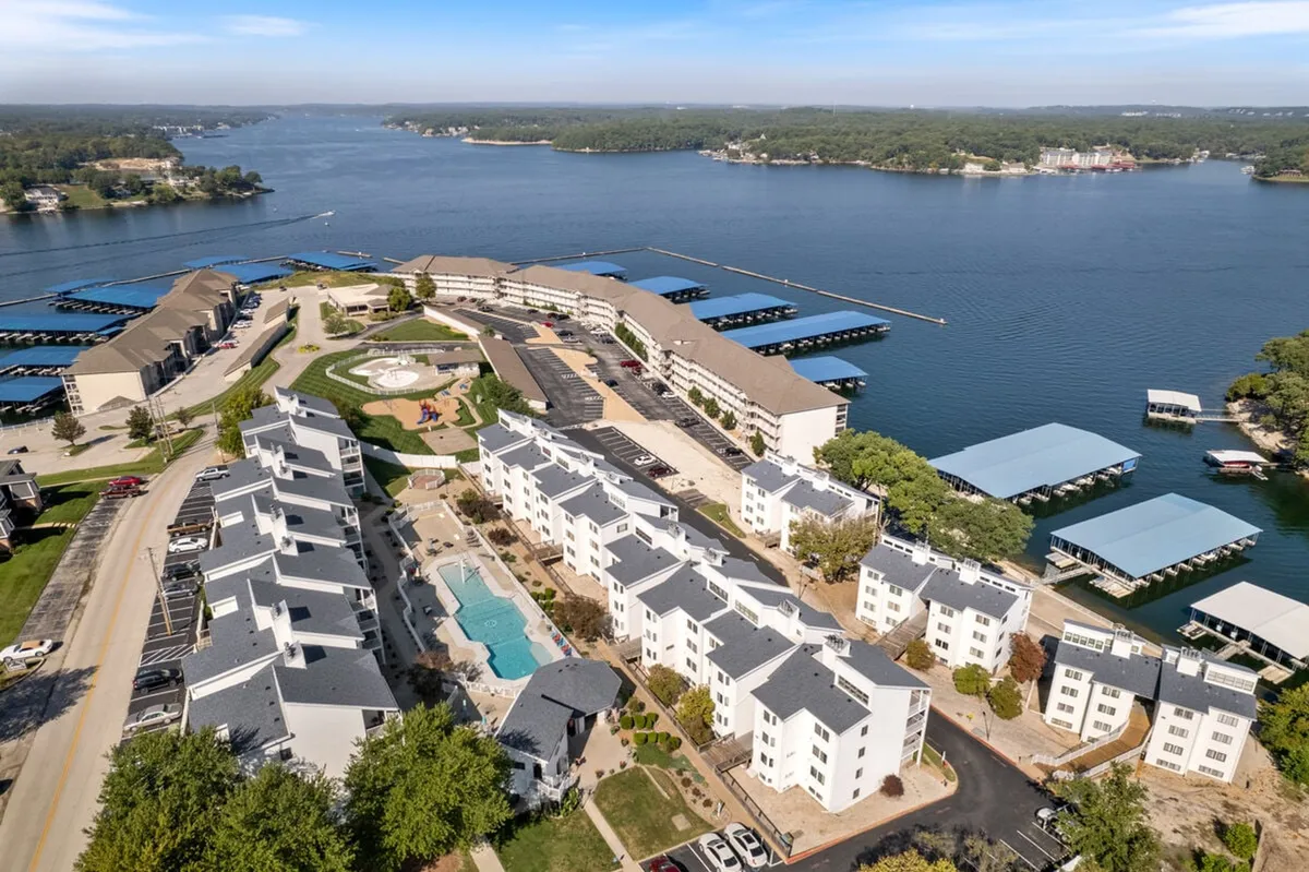 A vibrant exterior shot capturing a sparkling swimming pool surrounded by residential buildings. The scene includes a playground, glimpses of a lake, and boat docks, with people potentially enjoying various amenities.