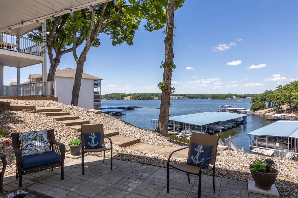 A peaceful lakeside scene featuring a seating area overlooking the water, with a dock and boats visible in the background. The view includes both the serene lake and comfortable outdoor furniture, ideal for relaxing or enjoying a drink with panoramic water views.