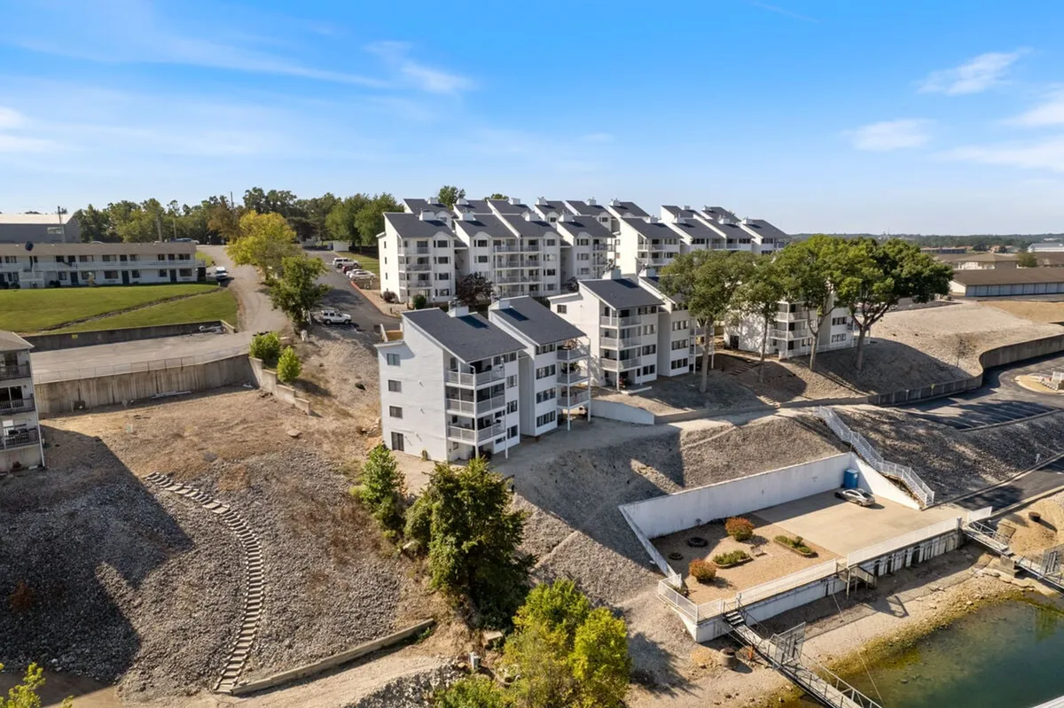 A vibrant exterior view of modern apartment buildings situated right on the lakefront, with landscaped gardens, walking paths, and a visible waterfront area. Children can be seen playing in a nearby park, and the setting sun casts a warm glow over the community.